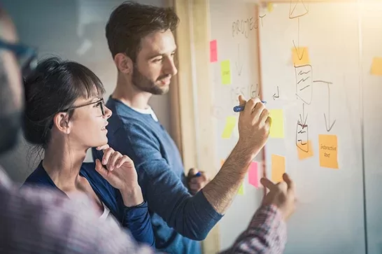 Duas pessoas em frente a um quadro de vidro com post-its coloridos, escrevendo e discutindo um diagrama durante uma sessão de brainstorming.