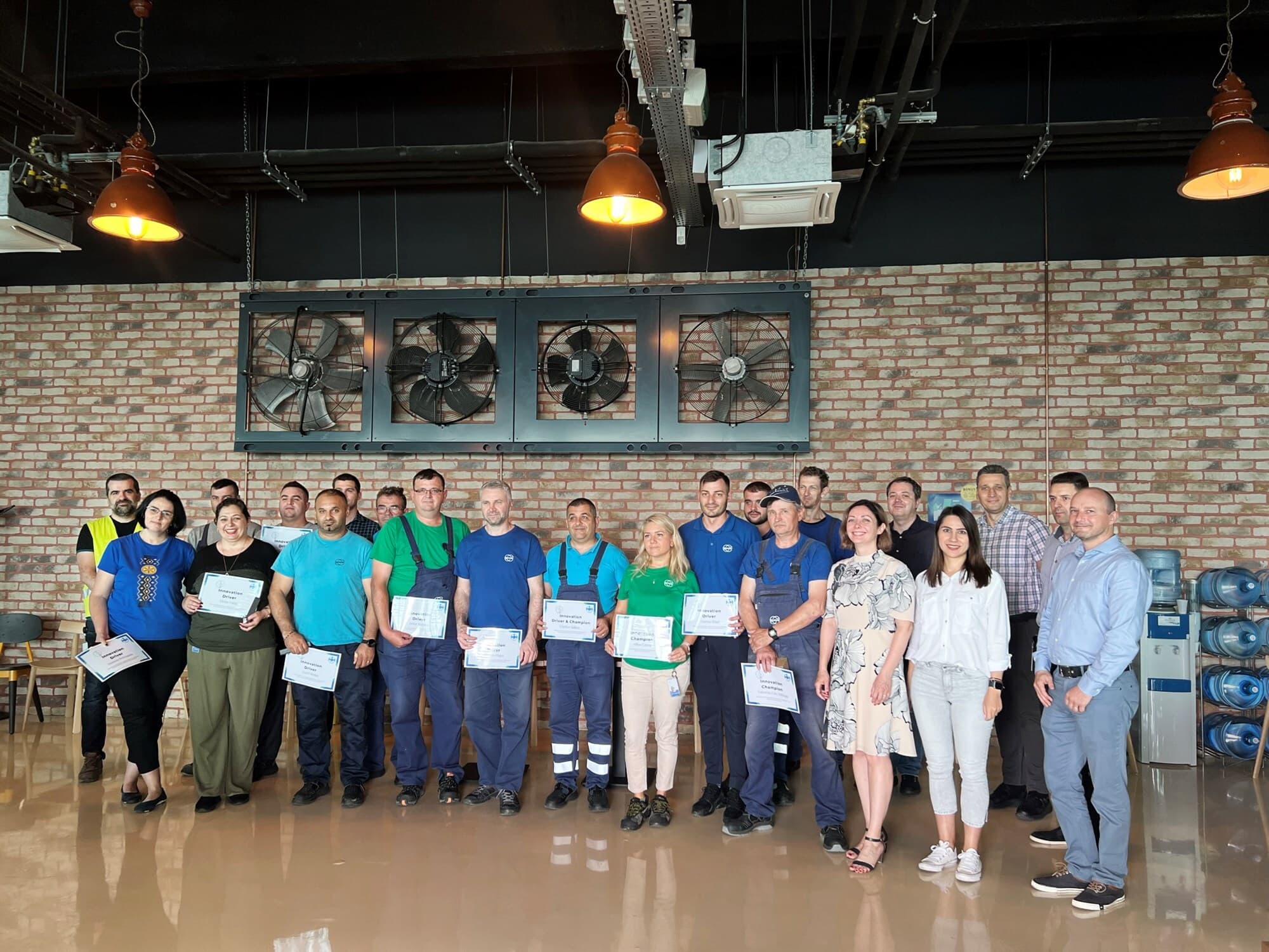 A diverse group stands in an industrial-style room with brick walls, large wall fans, and overhead orange pendant lights, several people holding certificates.