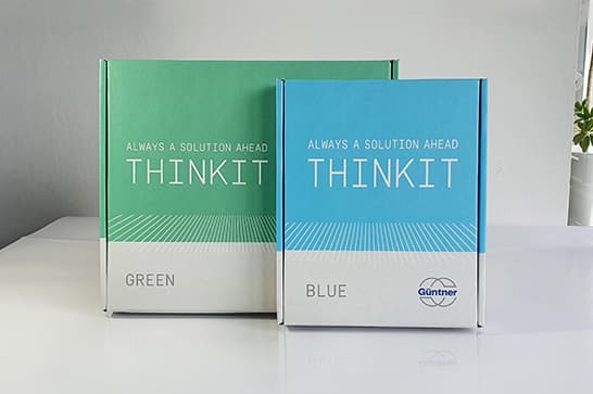 Two Thinkit product boxes in green and blue on a white surface, featuring 'Always a solution ahead' and 'GREEN'/'BLUE' labels; plant visible at the edge.