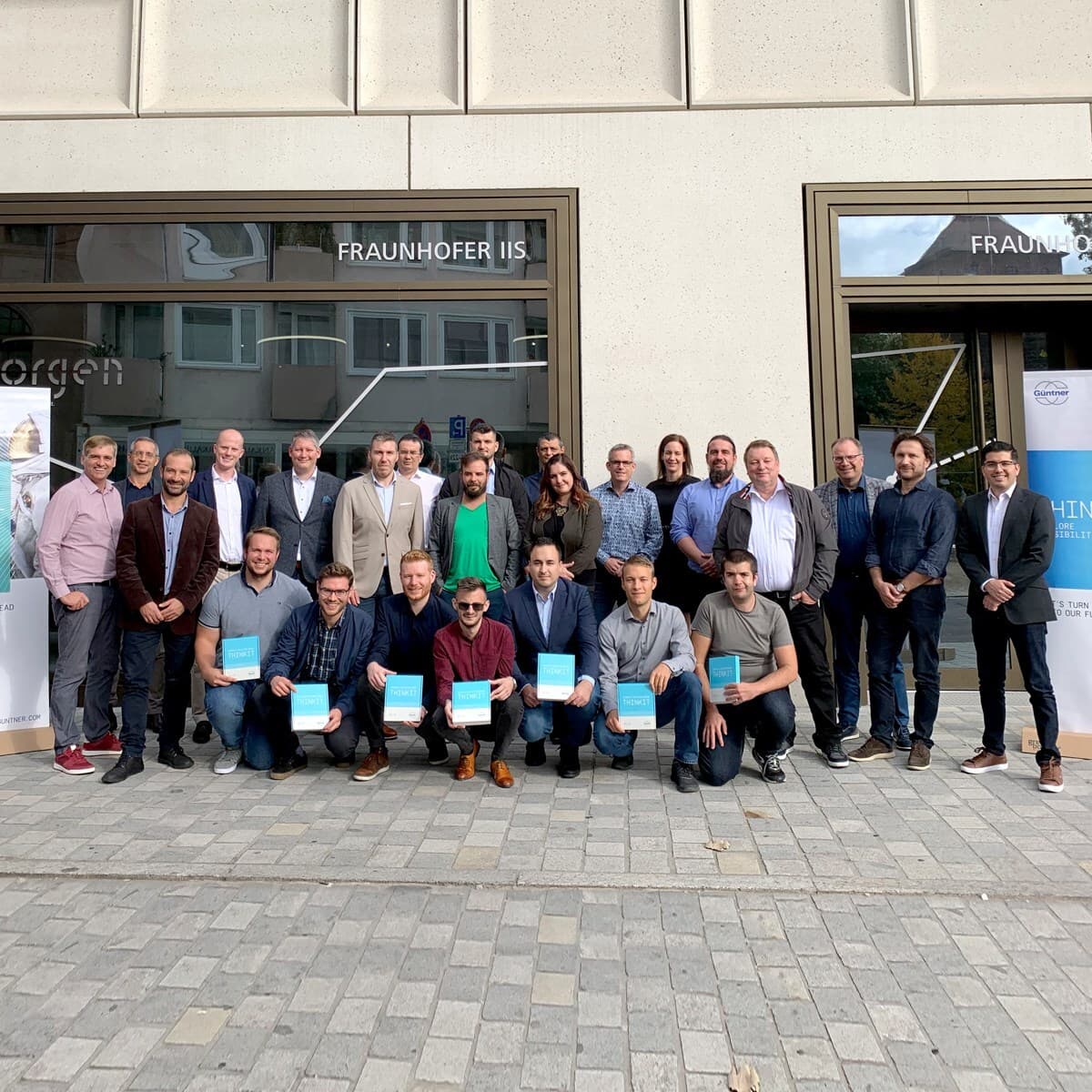 Group of diverse professionals posing outside a modern building with FRAUNHOFER IIS signage; several hold blue certificates.
