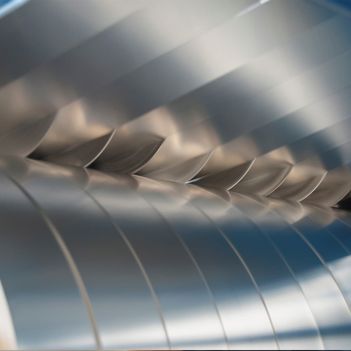 Close-up of shiny, curved metal blades in a diagonal row, resembling a turbine, with a soft blue-gray background.