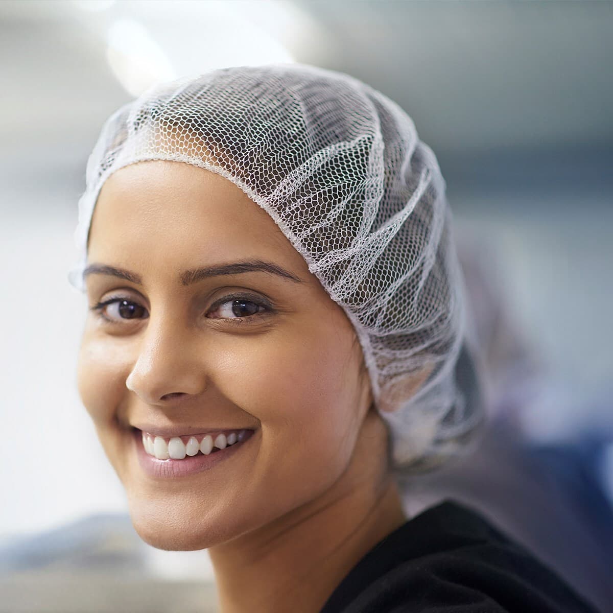 Close-up of a smiling person wearing a white mesh hairnet in a clinical or lab setting.