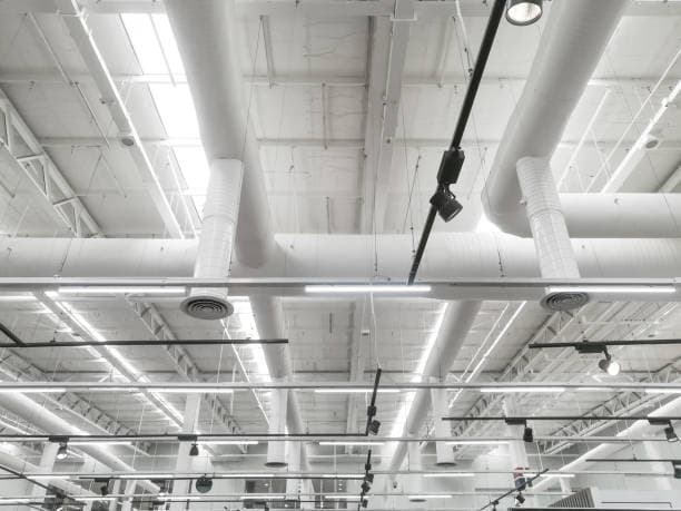 Industrial ceiling interior with exposed white pipes, ducts, and track lighting across a bright, airy space.