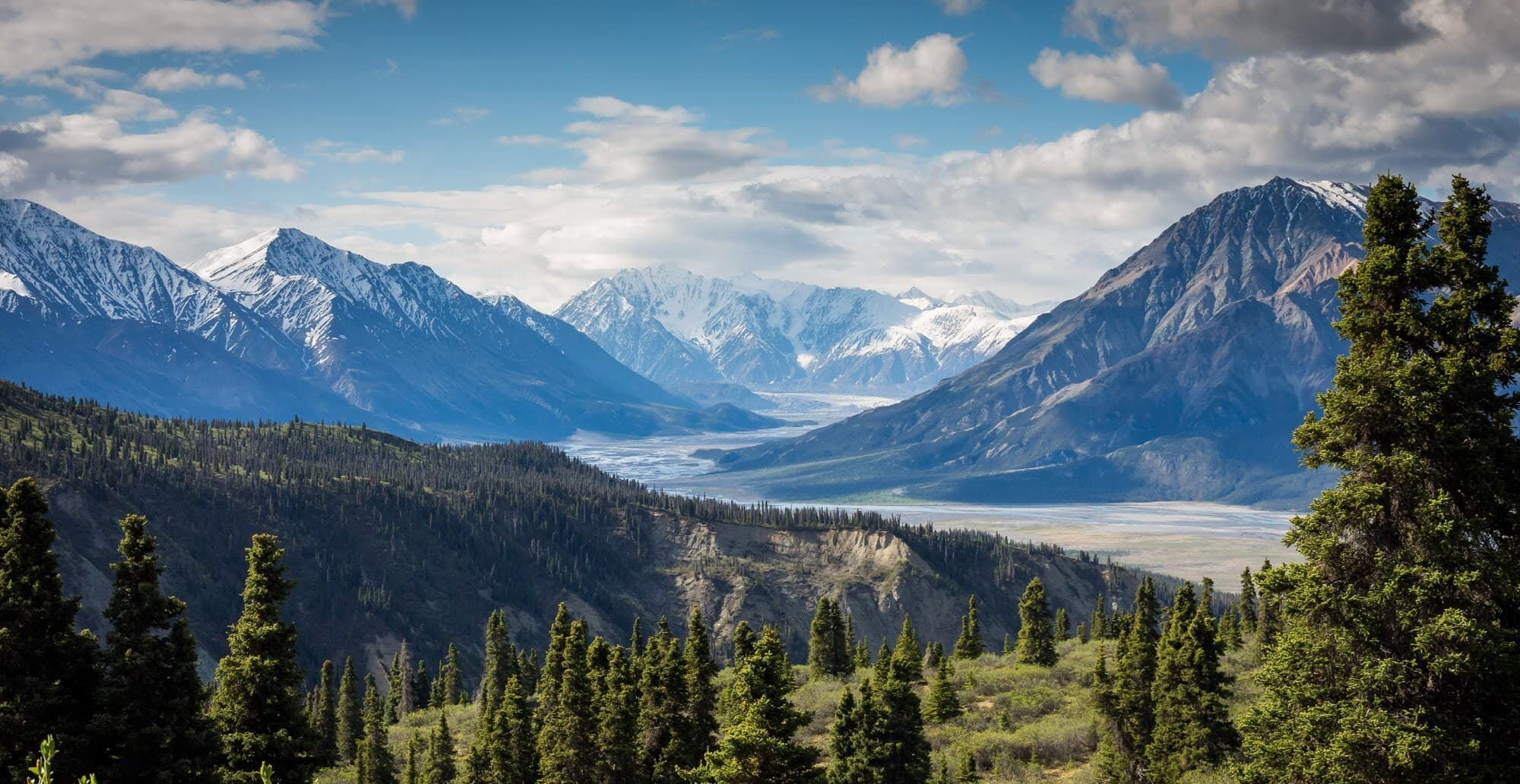 Snow-capped mountains tower over a green valley with a distant river, evergreens in the foreground, under a blue sky with scattered clouds.