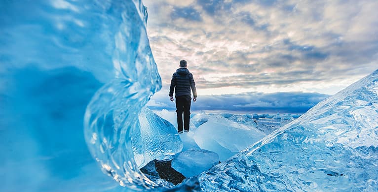 Una persona de espaldas, con chaqueta oscura, de pie entre bloques de hielo azul en un glaciar, mirando el cielo nublado.