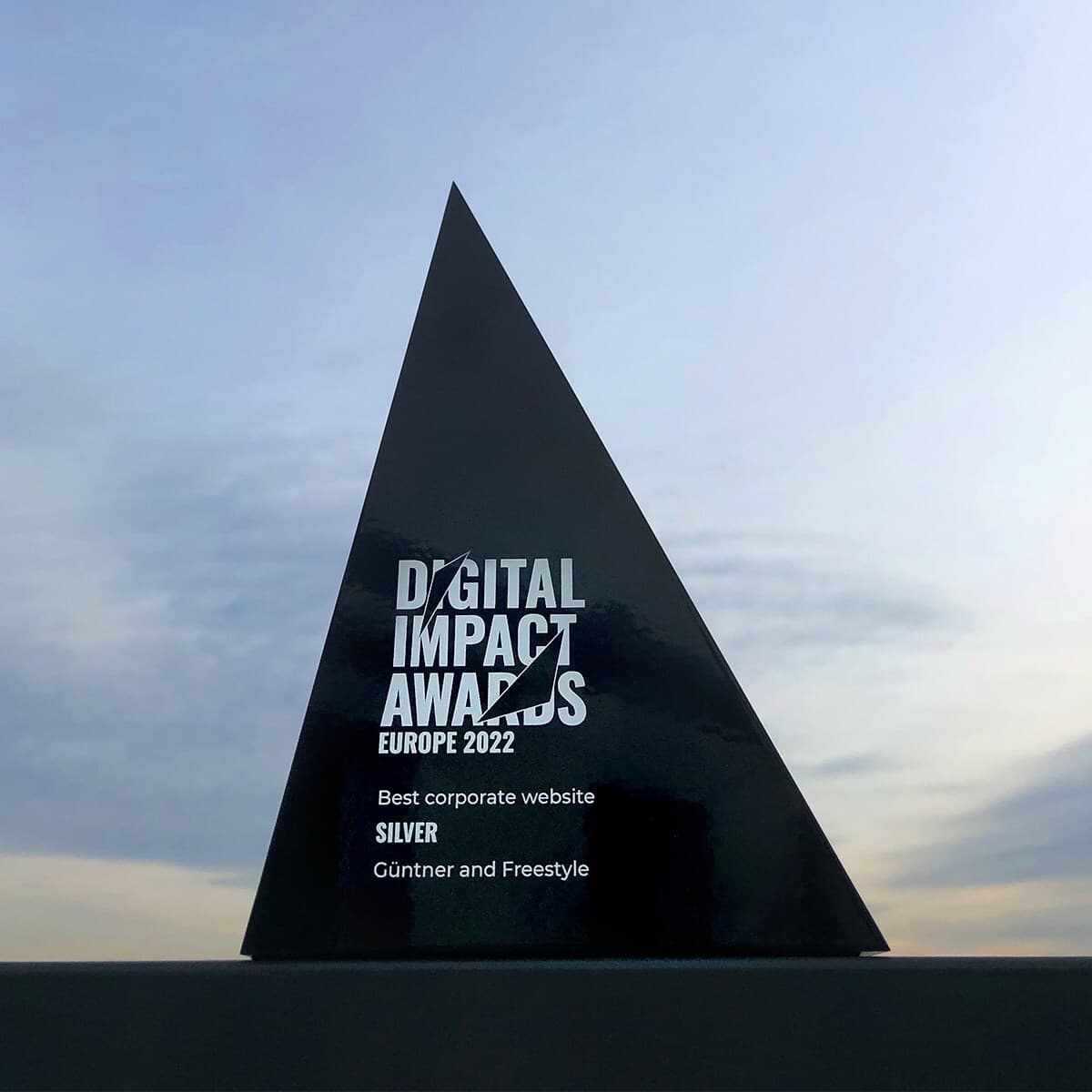 Tall black triangular award plaque on a ledge, with white text reading "Digital Impact Awards Europe 2022" and "Best corporate website SILVER Günther and Freestyle" against a pale sky.