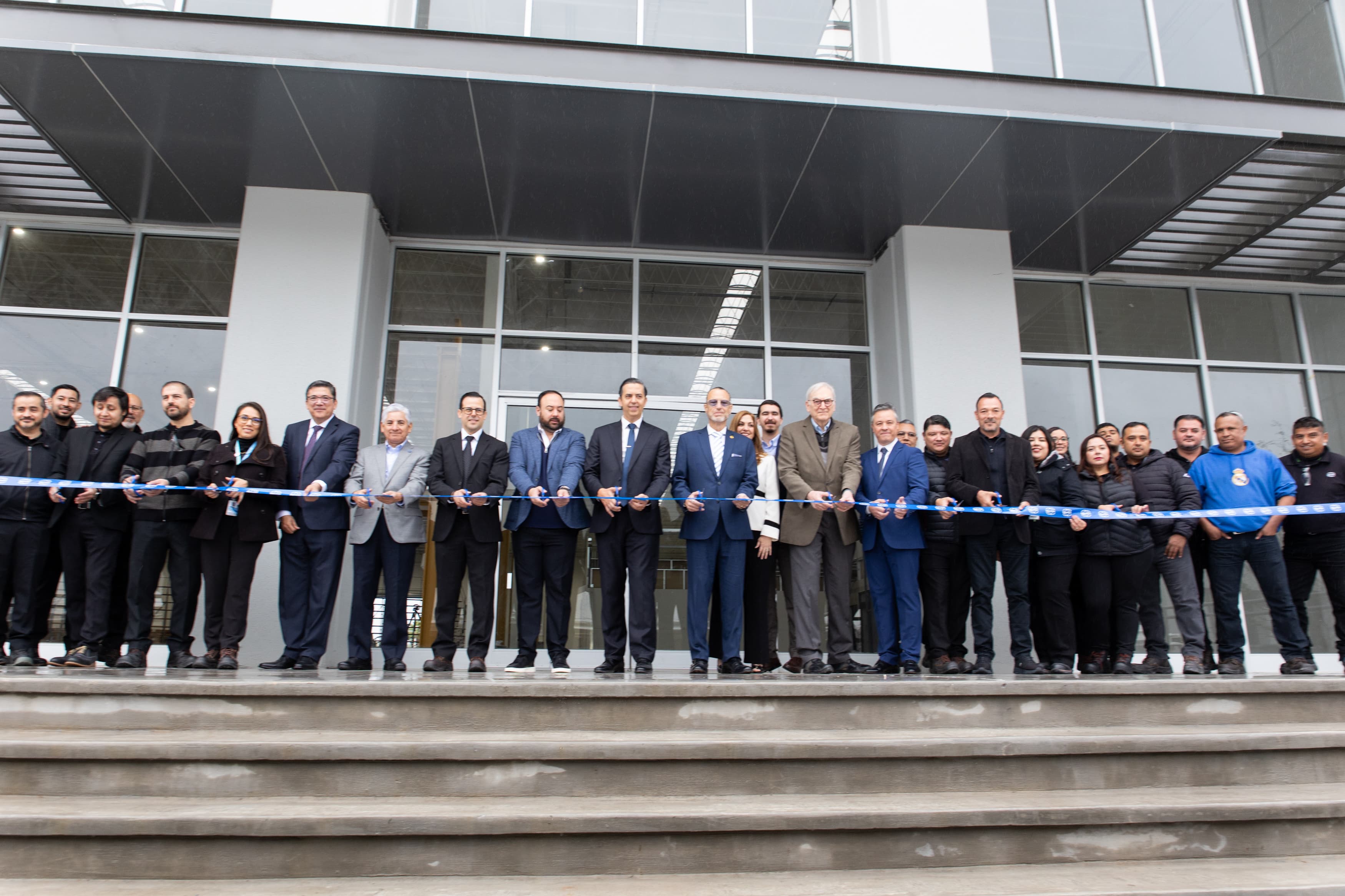 Group of people in business attire stand on steps in front of a modern glass building, holding a blue ribbon for a ceremonial ribbon-cutting.