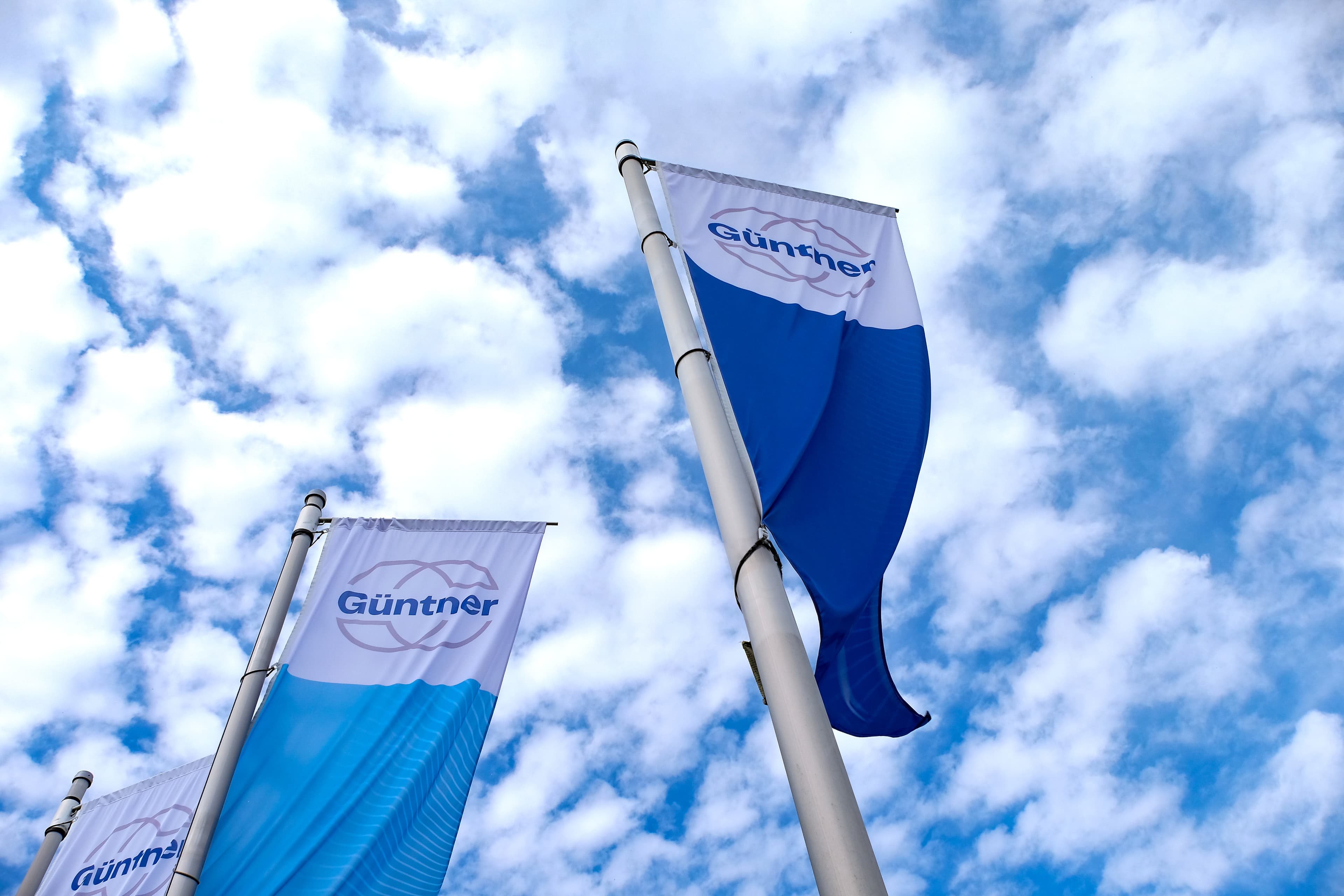 Trois bannières bleu et blanc affichant « Günther » qui flottent sur des mâts au vent, sous un ciel bleu avec des nuages blancs.