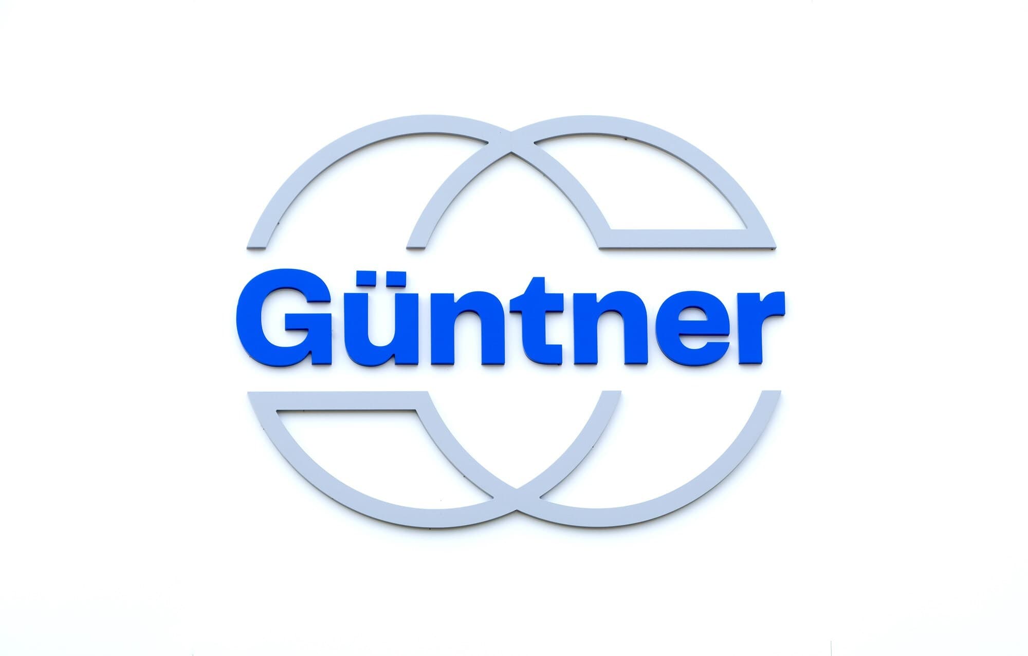 Logo avec le texte Günther en bleu gras, entouré de deux arcs gris qui se croisent pour former un cercle sur fond blanc.