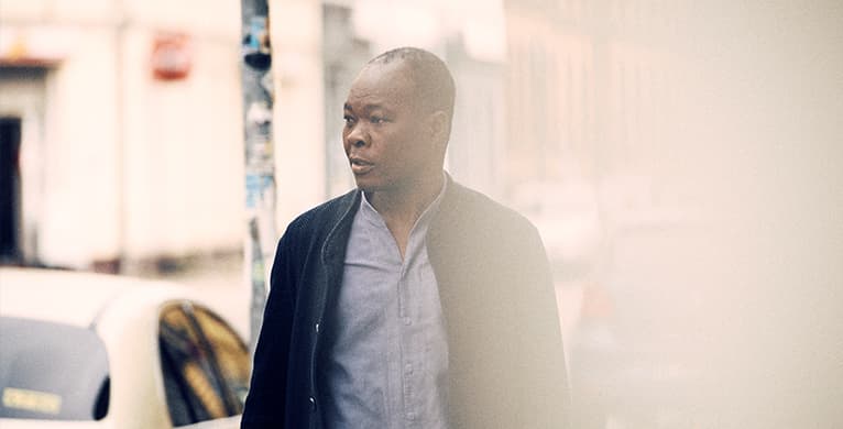 A person walks along a city street, wearing a light blue shirt and dark jacket; background is blurred with warm, hazy light.