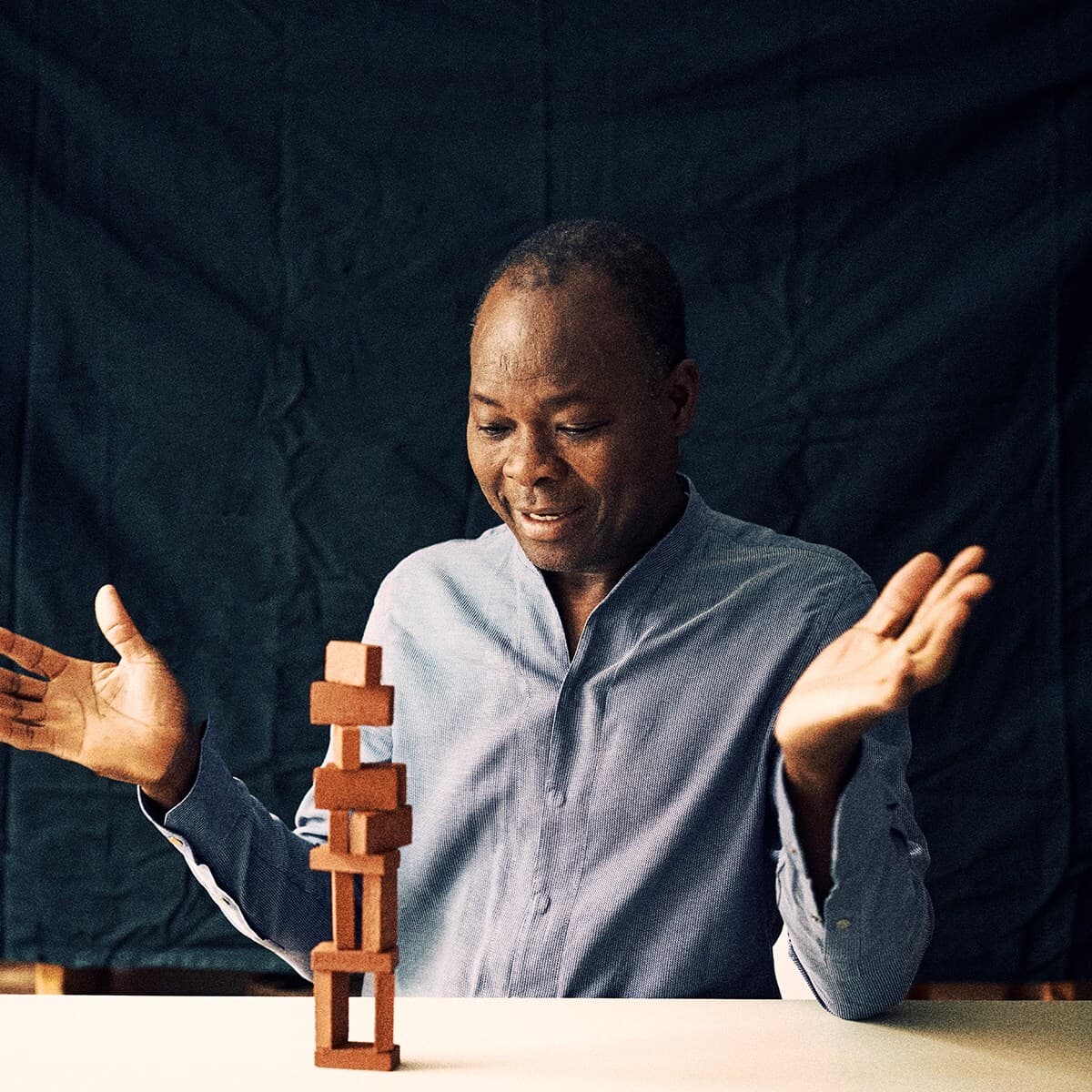 A person in a light blue shirt sits at a white table, with a small smile and hands raised, while a wooden block tower stands in front of them.