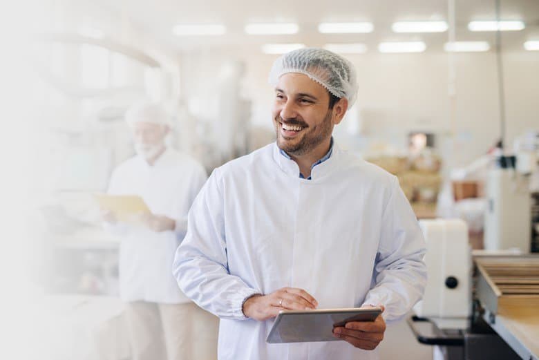 Smiling person in a white lab coat and hairnet holds a tablet in a bright, clean production facility with blurred colleagues in the background.