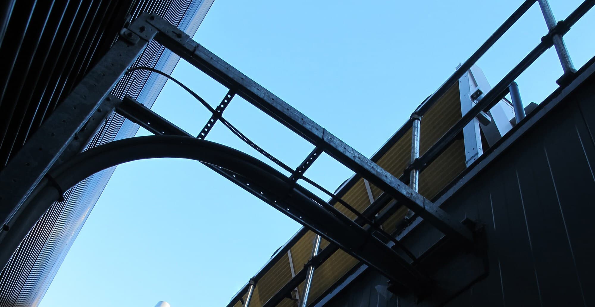 Low-angle view of steel framework, pipes, and railing attached to a building, with a clear blue sky in the background.