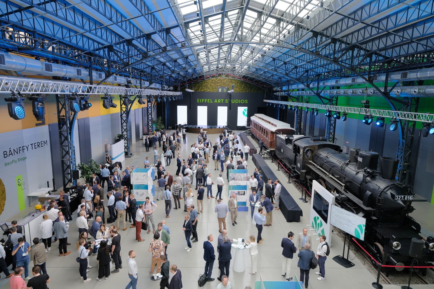 Busy indoor rail hall expo; attendees mingle at booths as a vintage steam locomotive and train cars sit to the right under blue steel arches.