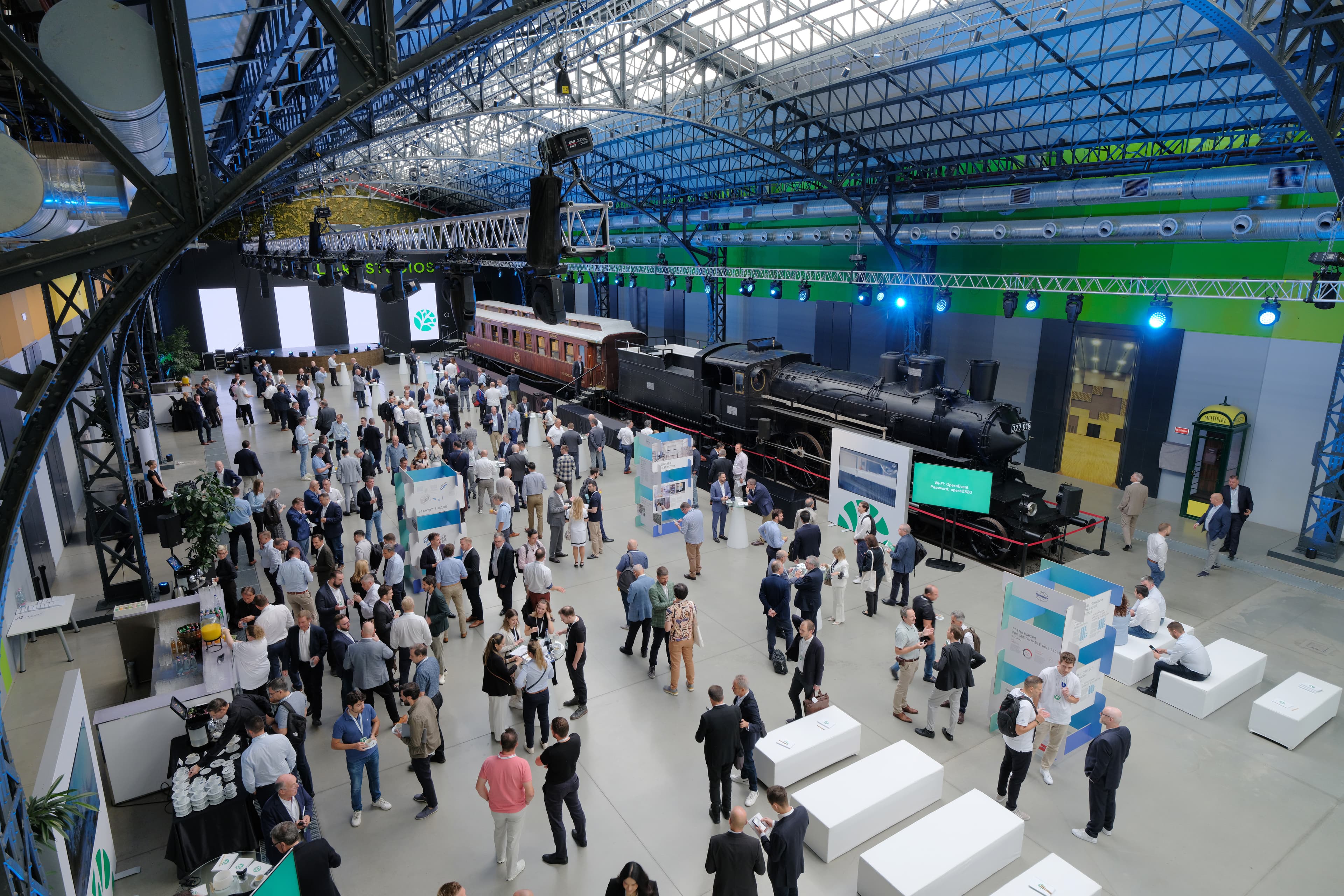 A large indoor exhibition hall with a steam locomotive and a vintage passenger carriage on display, as groups of attendees mingle near booths and screens.