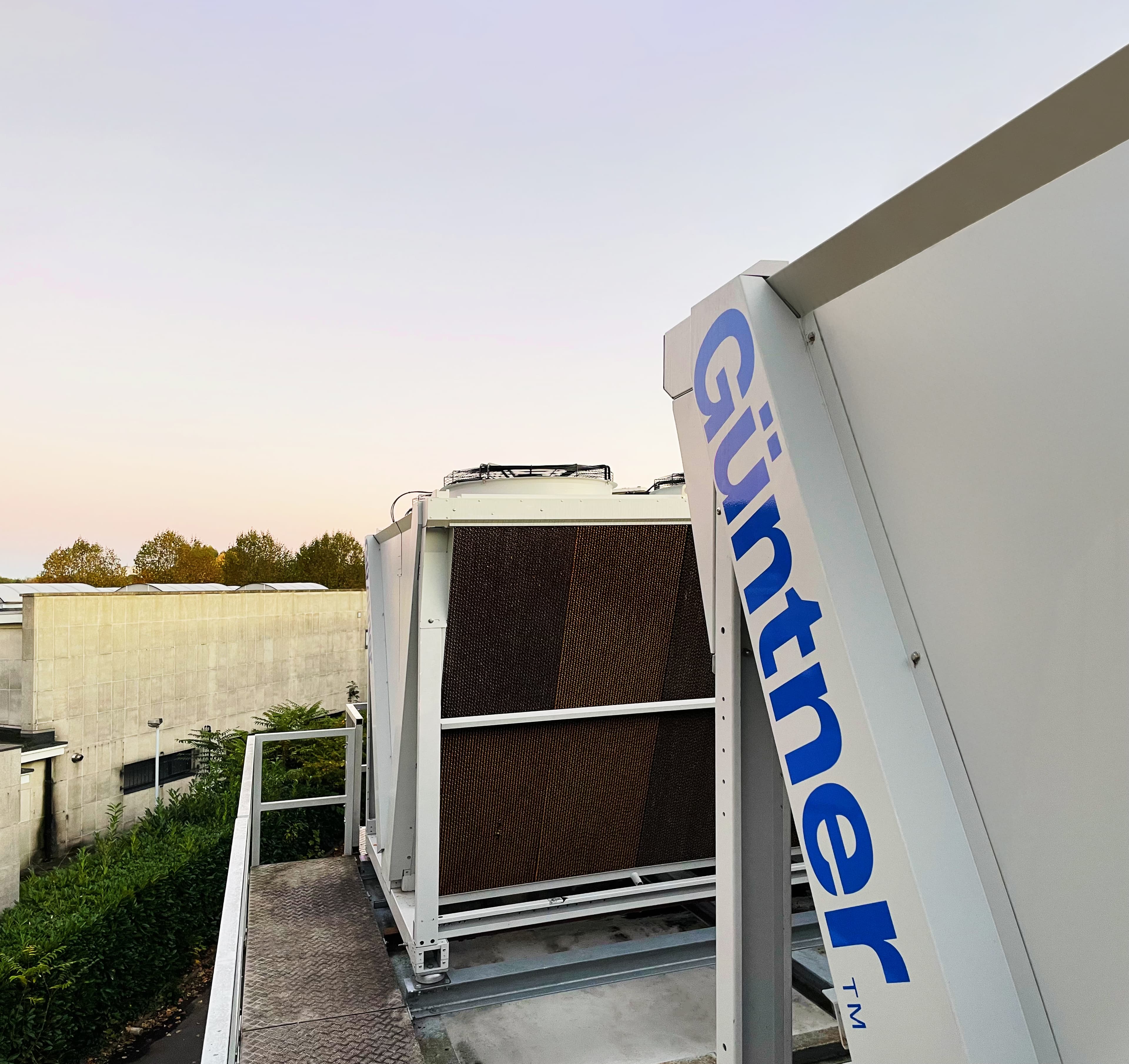 Rooftop HVAC unit with a large heat exchanger, white panel bearing a blue 'Guntner' logo, against a pastel sunset sky and distant trees.