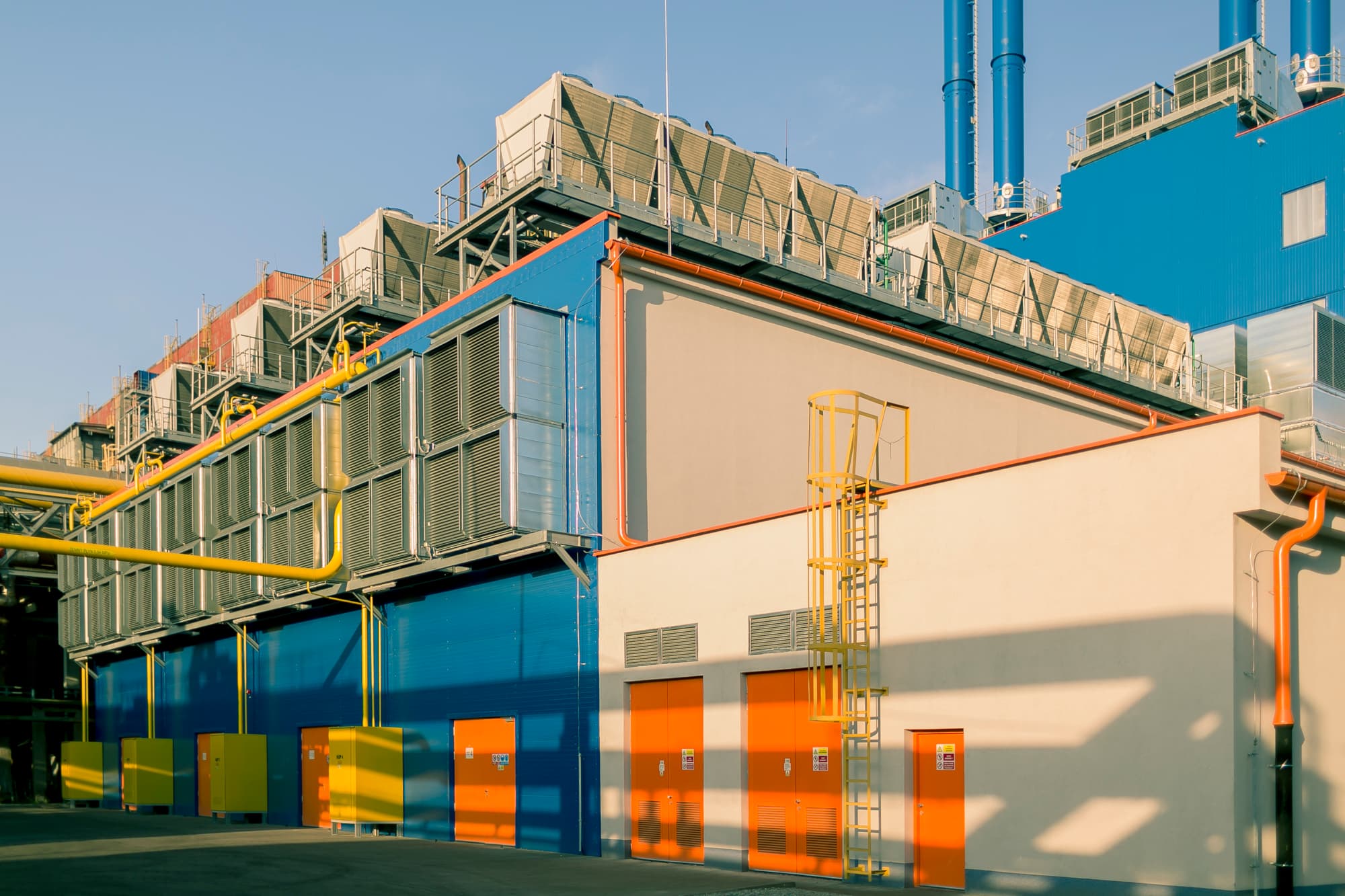 Industrial facility with blue and orange exterior, row of vented units along the wall, yellow pipes, a tall yellow ladder, and a clear blue sky.