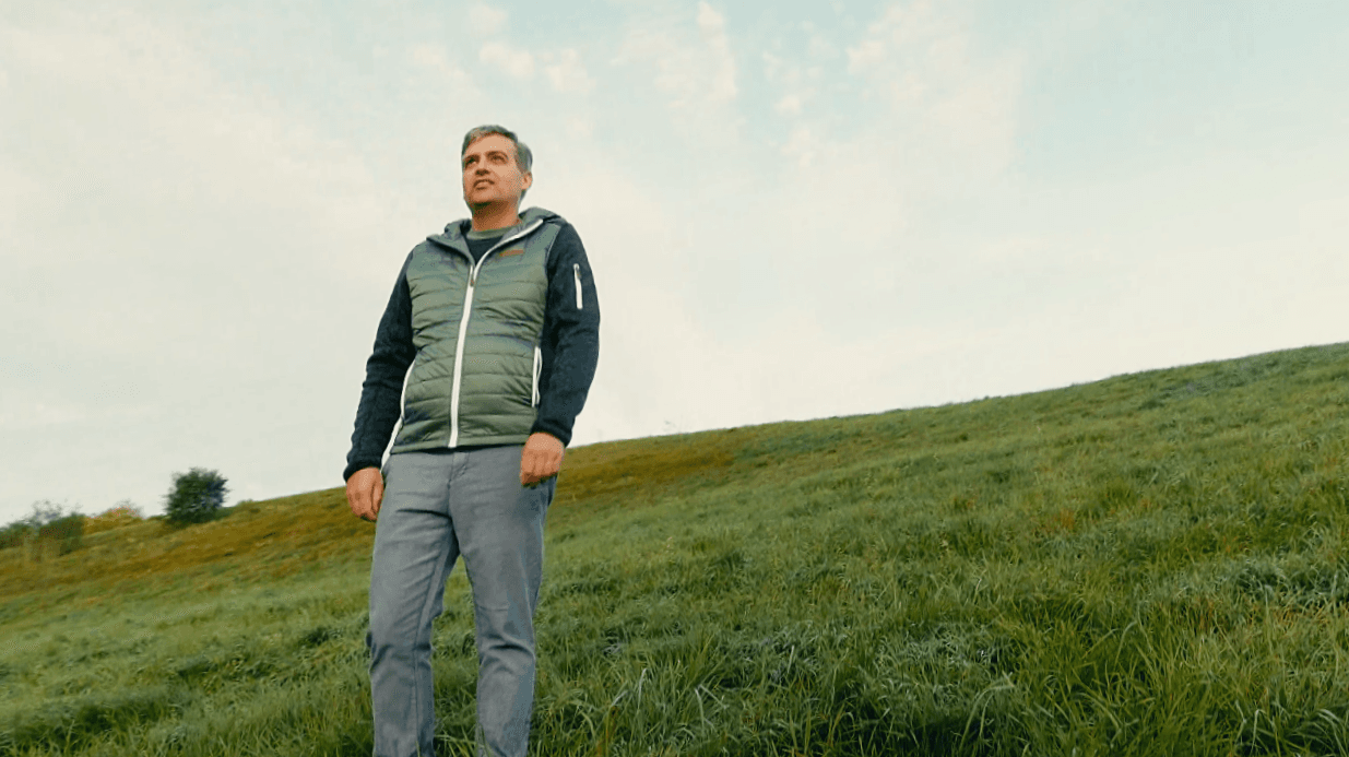 A person stands on a grassy hillside, wearing a green puffer vest and jeans, looking up at a pale blue sky.