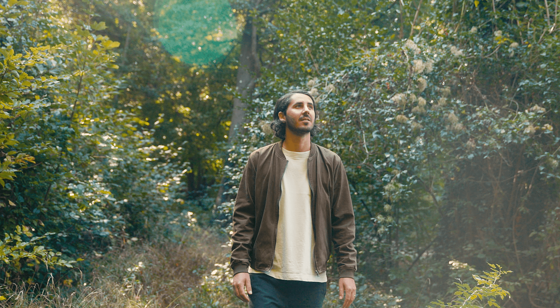 A person with dark hair wearing a brown jacket over a light shirt stands in a sunlit green forest, looking upward.