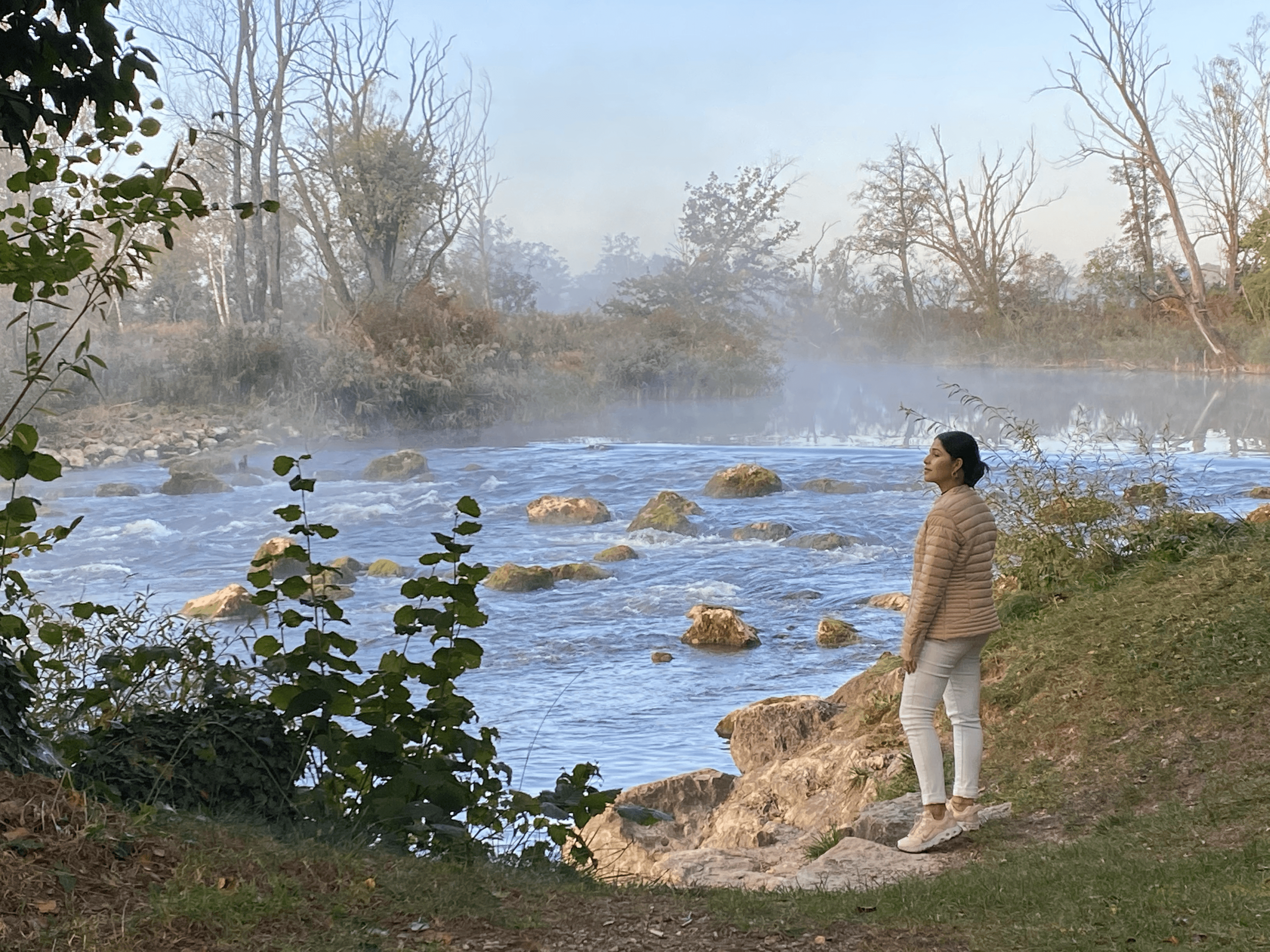 Person standing on a rocky riverbank in a beige quilted jacket and white pants, gazing at a misty river with rocks and bare trees.