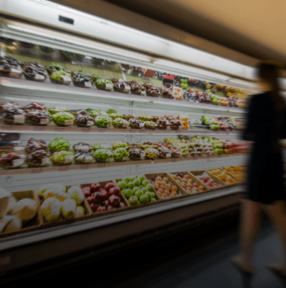 Blurred grocery store produce aisle with colorful fruits and vegetables on shelves; a person stands on the right, slightly out of focus.