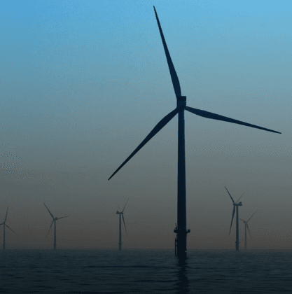 Offshore wind turbine in the foreground over calm water, with several distant turbines on a hazy horizon under a blue-to-orange gradient sky.
