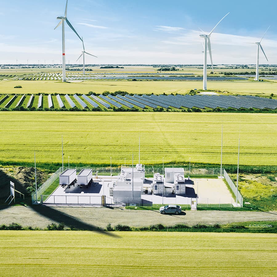 Aerial view of a rural energy site: rows of solar panels, several wind turbines on the horizon, and a fenced utility facility with a car nearby.