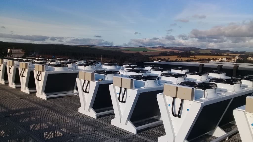 Rows of white industrial cooling units with fans on top sit on a rooftop, with a countryside landscape of fields and hills beyond.