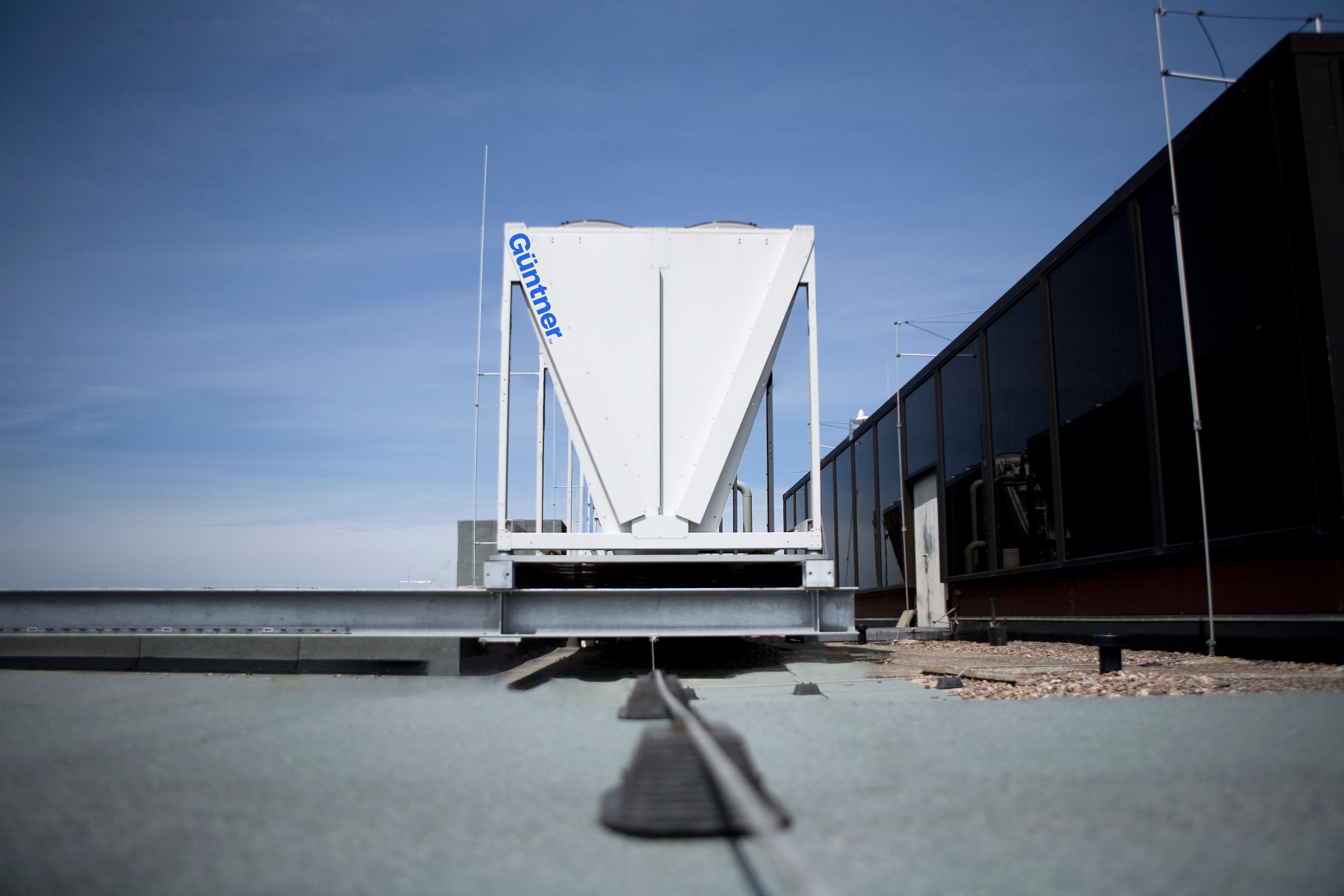 Rooftop with a white funnel-shaped industrial unit on a metal frame, a rail track in the foreground, and a dark glass building to the right under a blue sky.
