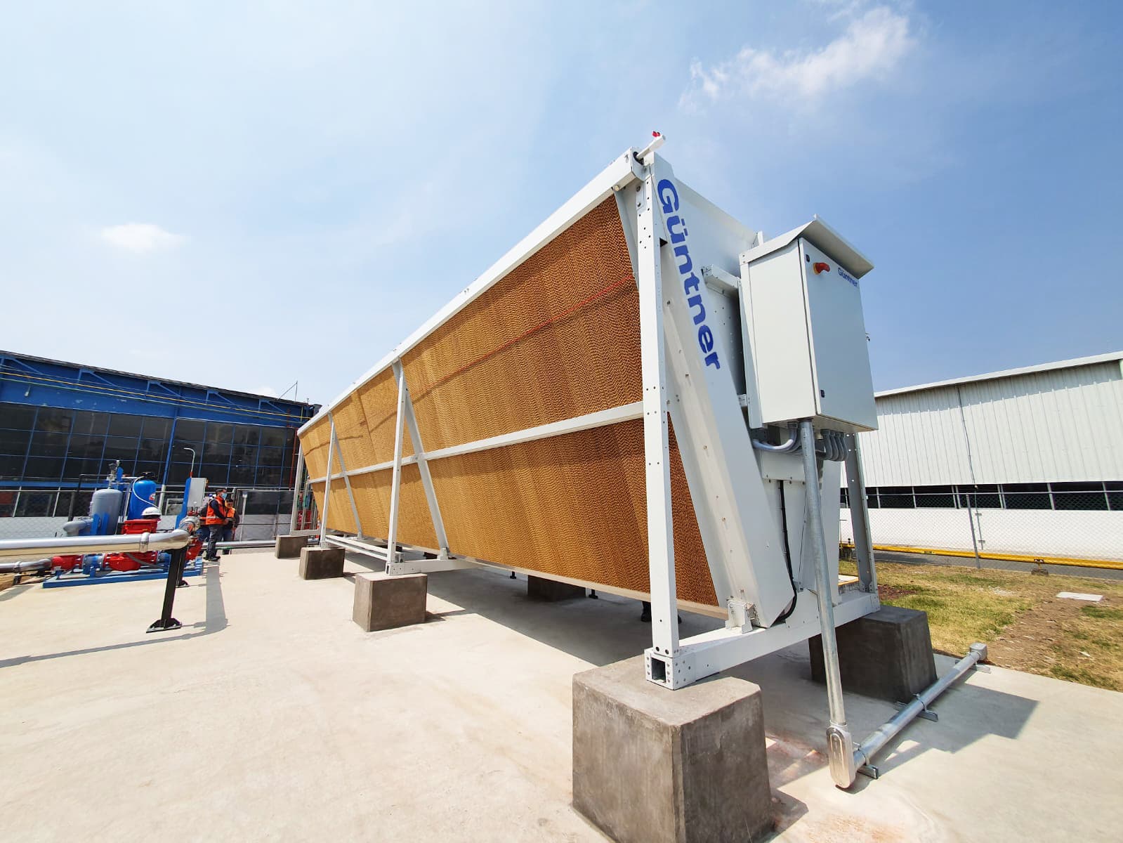 Guntner heat exchanger on a white frame with orange fins, mounted on concrete blocks; nearby pipes and industrial equipment, blue sky, and a worker in an orange vest in background.