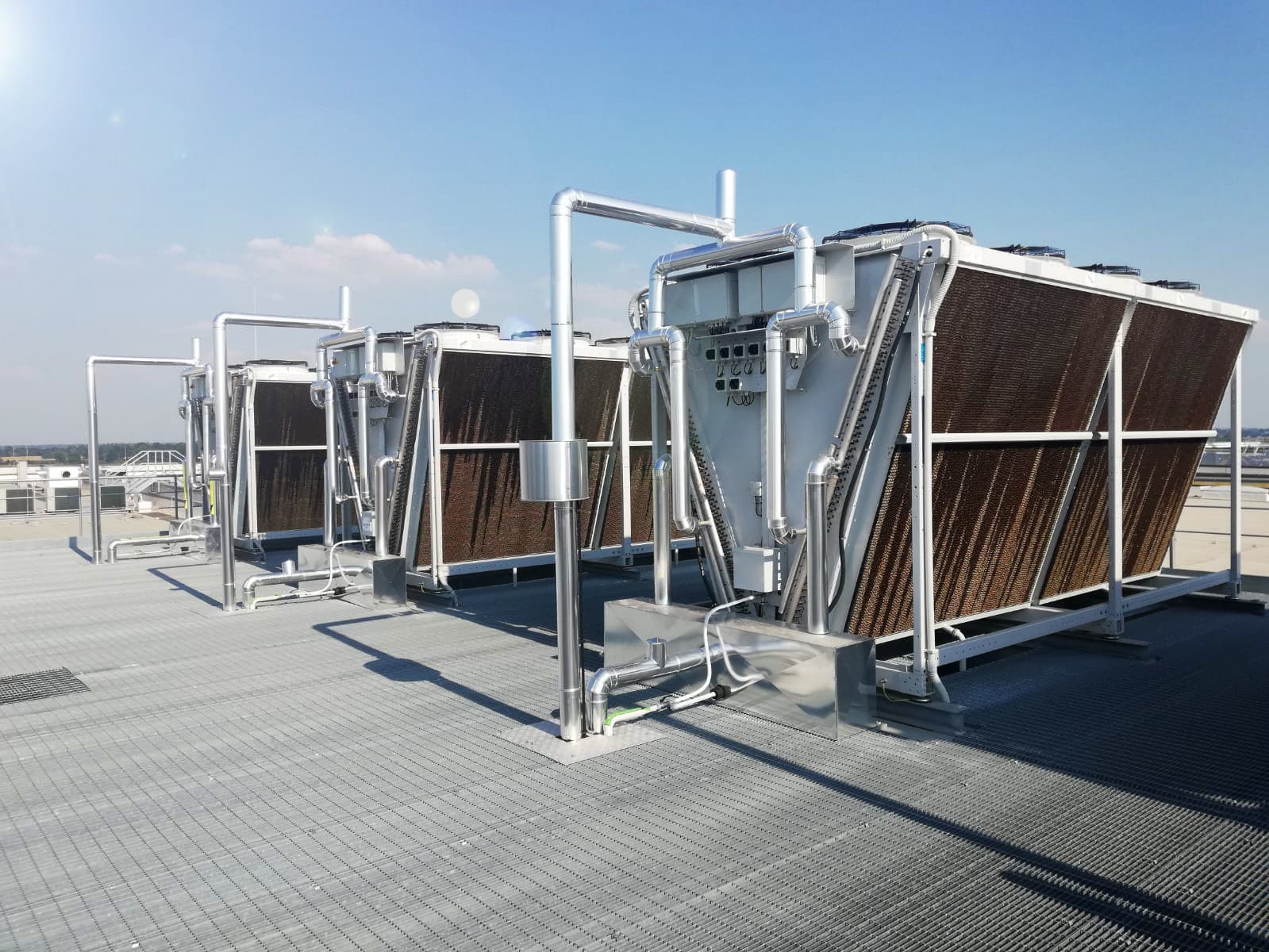 Rooftop scene with multiple large industrial cooling units framed in metal, interconnected silver pipes, and brown heat-exchanger fins under a clear blue sky.