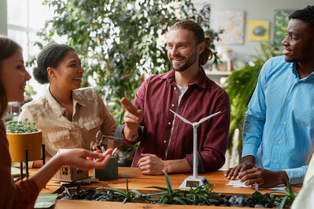 Four diverse people sit around a wooden table, smiling and discussing a small wind turbine model, with plants and bright indoor light in the background.