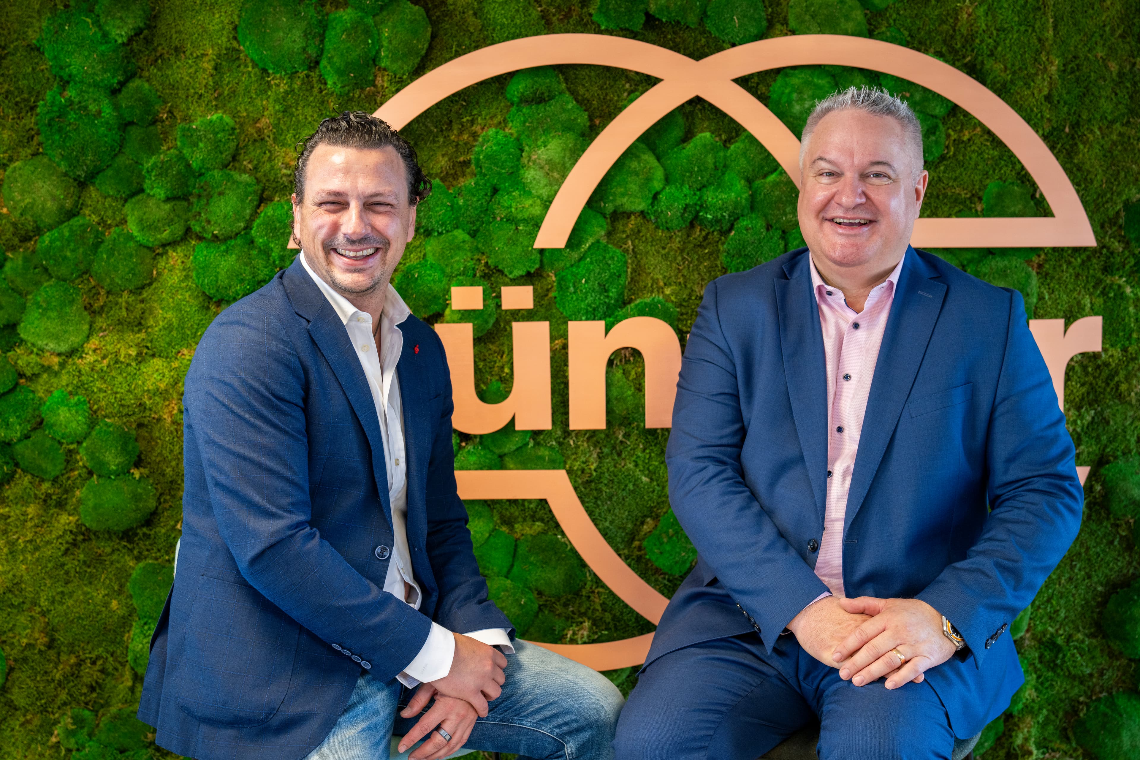 Two people in blue suits sit on stools before a green moss wall with a large orange logo, smiling at the camera.