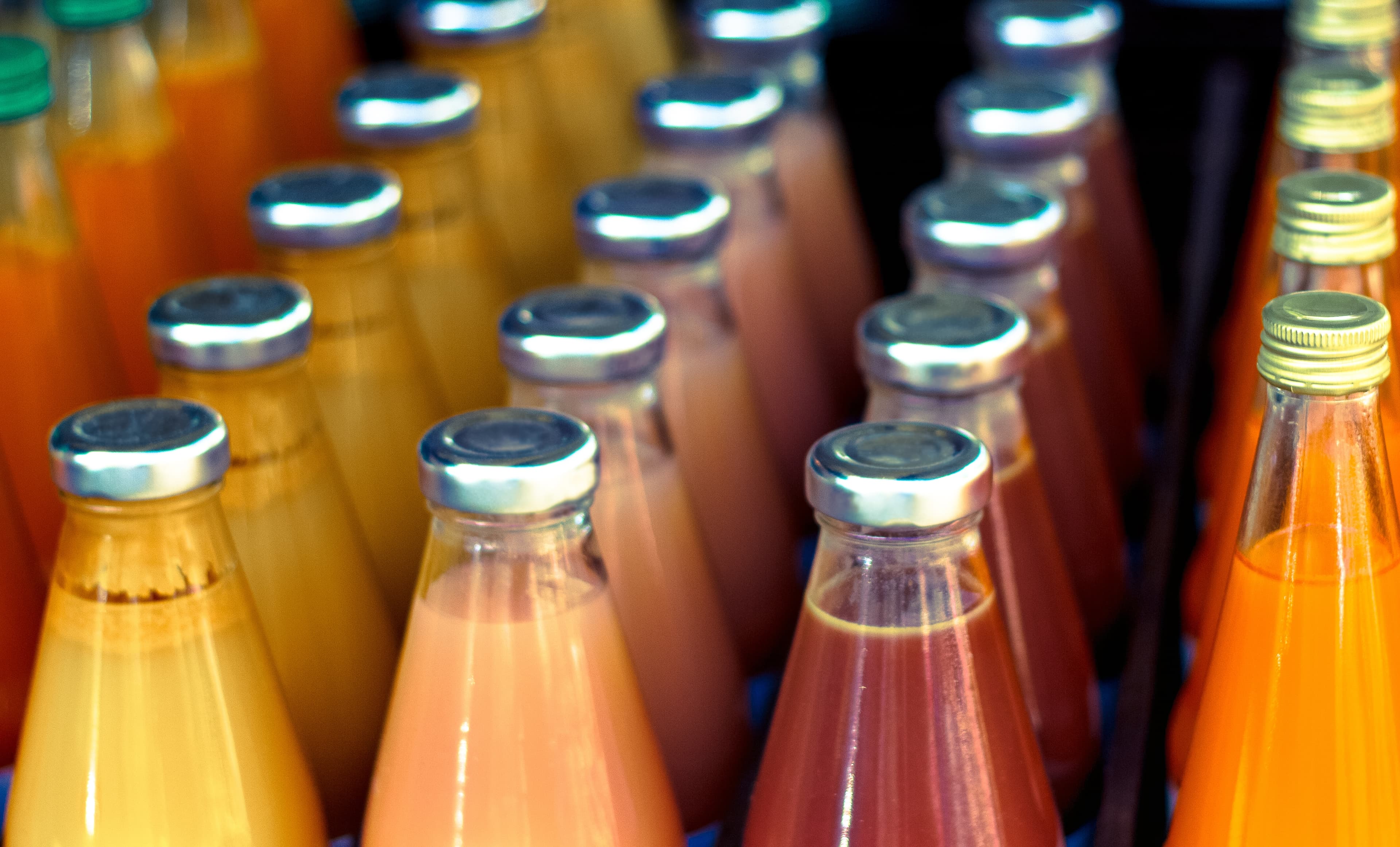 Close-up of rows of glass bottles with metal caps, filled with colorful beverages in orange, pink, and amber hues.