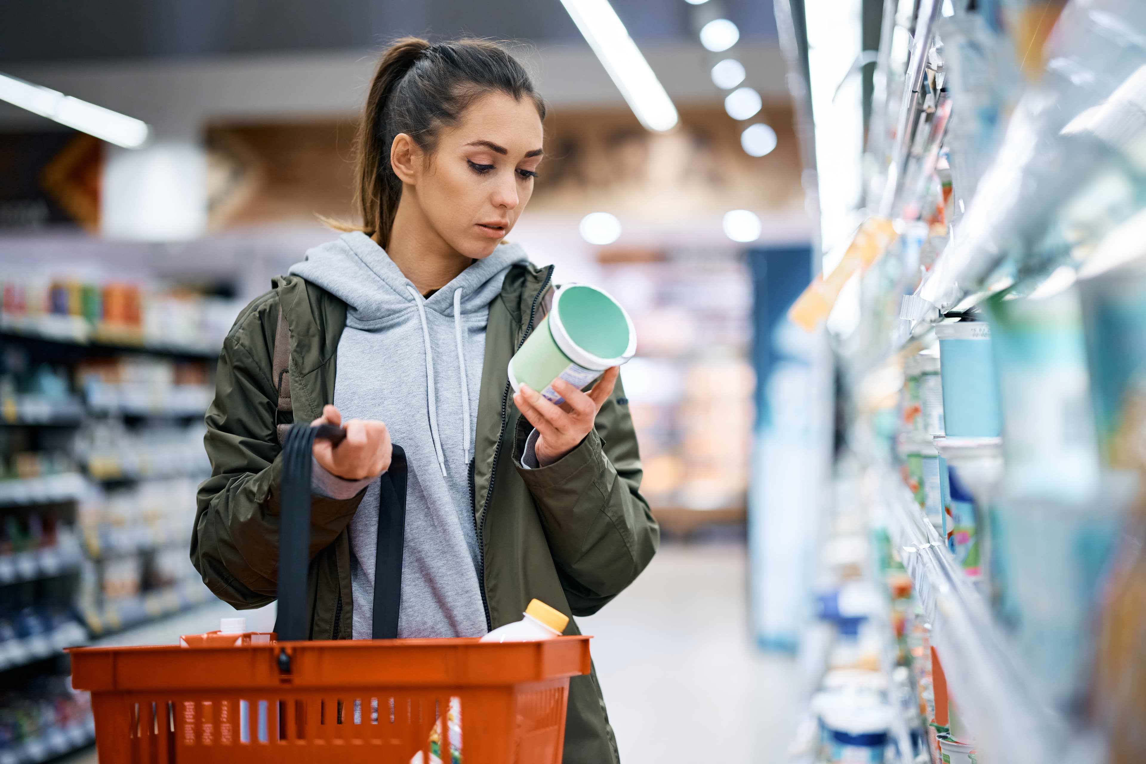 A shopper in a gray hoodie and green jacket examines a green-lidded container in a grocery aisle, with an orange basket in the foreground.
