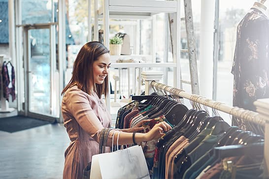 A person browses clothing on a rack inside a bright boutique, holding a shopping bag.
