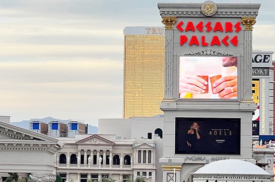 Caesars Palace marquee with red neon logo, a large screen showing hands with cards and a smaller screen featuring a performer, against Las Vegas hotel architecture.