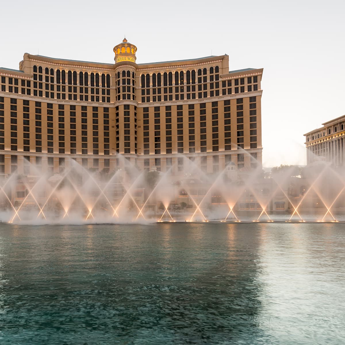 Bellagio hotel in Las Vegas with a curved façade and illuminated tower, fountains shoot arcing jets across the lake in front.
