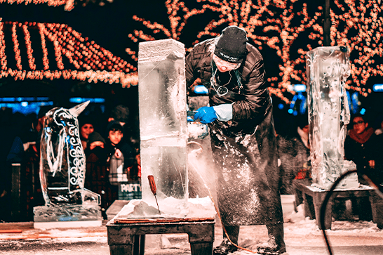 Ice sculptor wearing a mask and gloves chisels a block of ice on a wooden table, with festive string lights and spectators in the background.