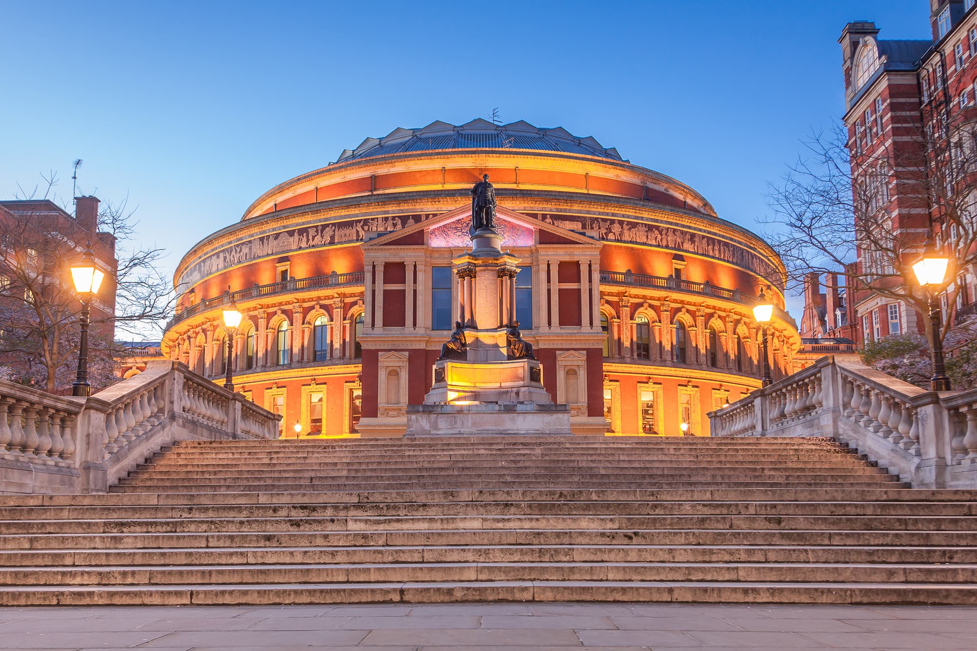 Dusk view of a round brick building lit in warm orange, with a central statue on a pedestal and wide stone steps flanked by railings.
