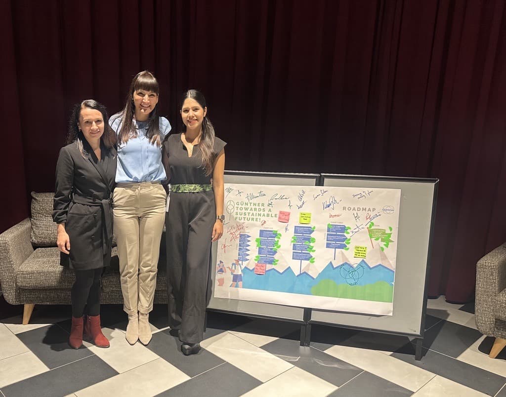 Three people stand next to a large poster with a colorful roadmap and signatures, in a room with a maroon curtain and gray seating.