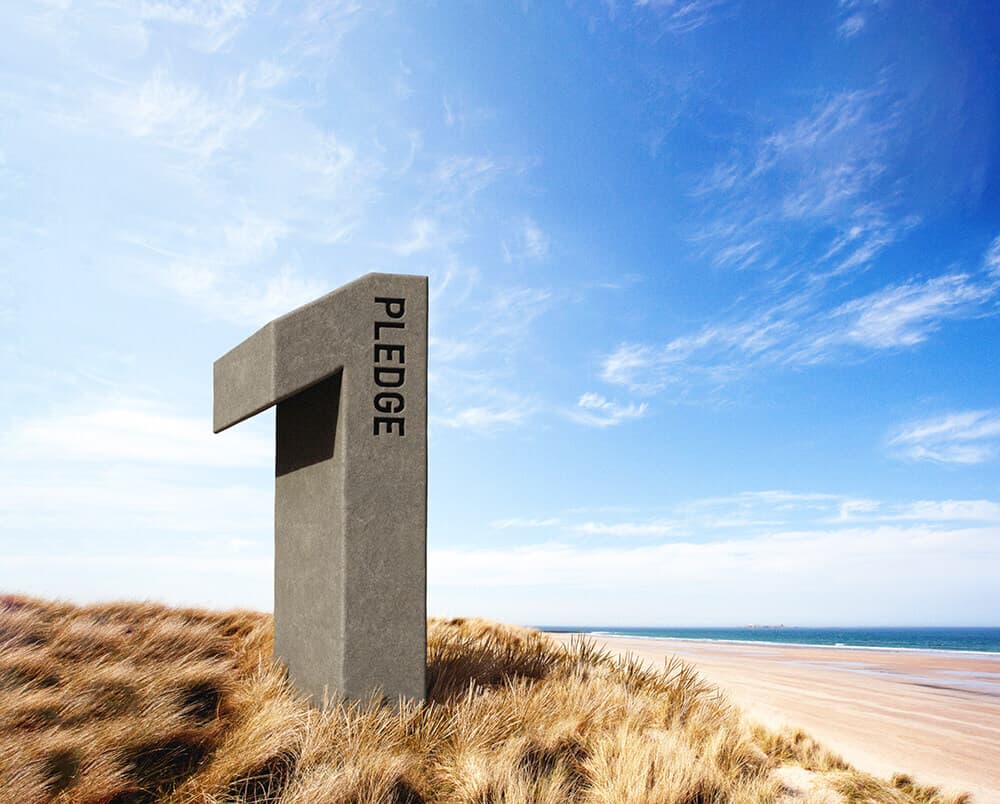 Large gray stone sculpture on a sandy dune, shaped like a bent pillar with PLEDGE written vertically, overlooking a beach and blue sea.