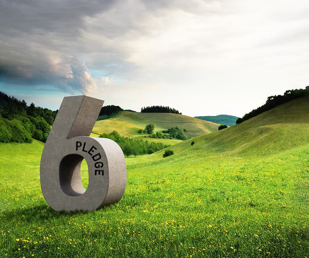Large gray stone sculpture of the number 6 with the word PLEDGE around its inner opening, in a green meadow with rolling hills and a cloudy sky.