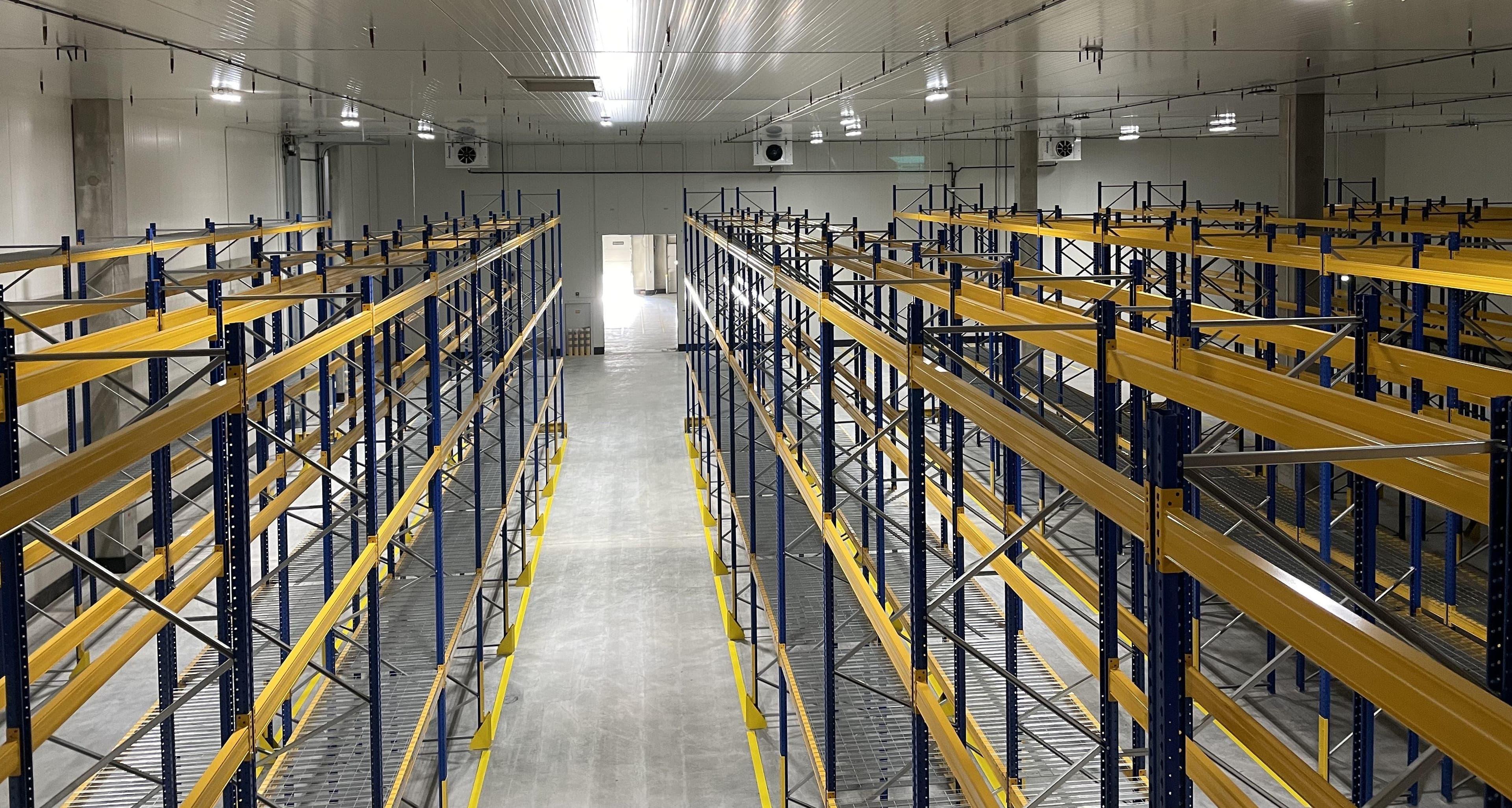 Industrial warehouse interior with tall blue and yellow pallet racks flanking a wide central concrete aisle under bright overhead lights.