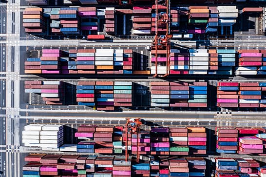 Aerial view of a busy shipping yard with rows of colorful stacked containers and large cranes lifting cargo overhead.