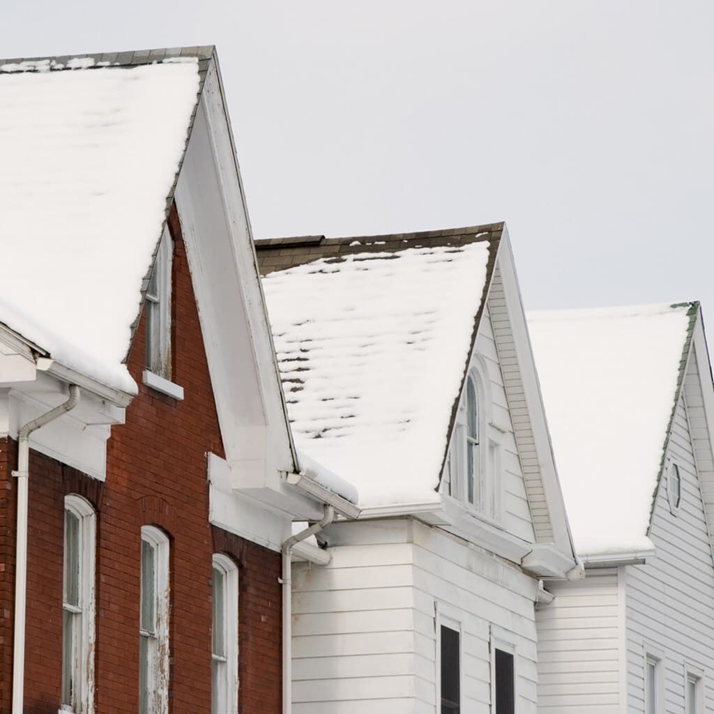 Snow-covered row of houses with steep gabled roofs; red brick and white siding in a wintry street scene.
