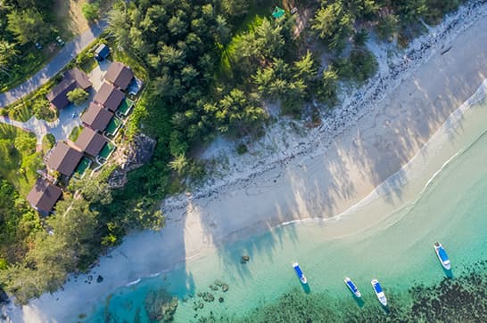 Aerial view of a white-sand beach with turquoise water, several small boats near shore, and a cluster of wooden cottages among pine trees beside a road.