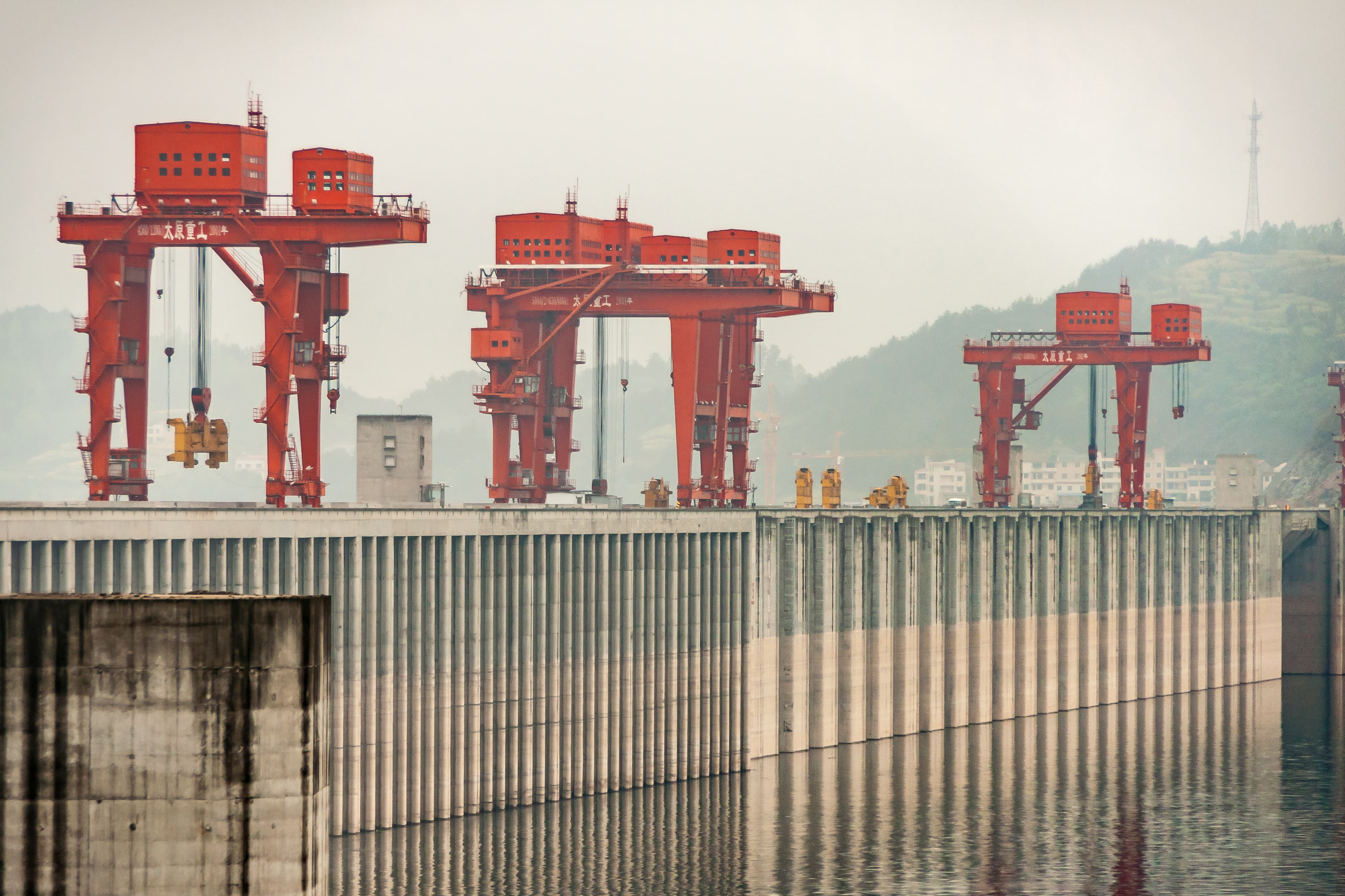 Several large red gantry cranes line the harbor, their hooks hanging over water, with a concrete seawall and misty hills in the distance.