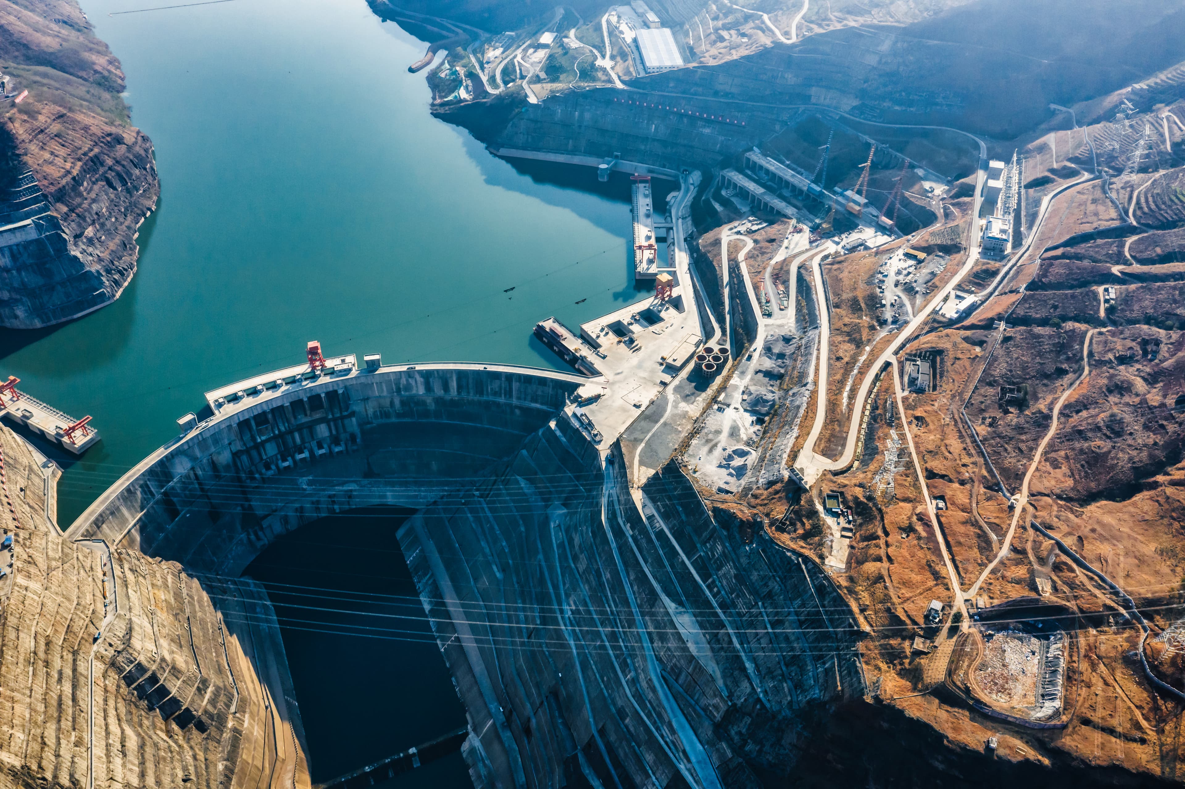 An aerial view of a curved concrete dam with a turquoise reservoir, spillways, and winding roads across brown, rugged hills.