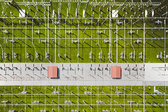 Aerial view of a grassy field with a grid of metal antenna towers; a light road crosses the center with two small red-roofed huts and long shadows.